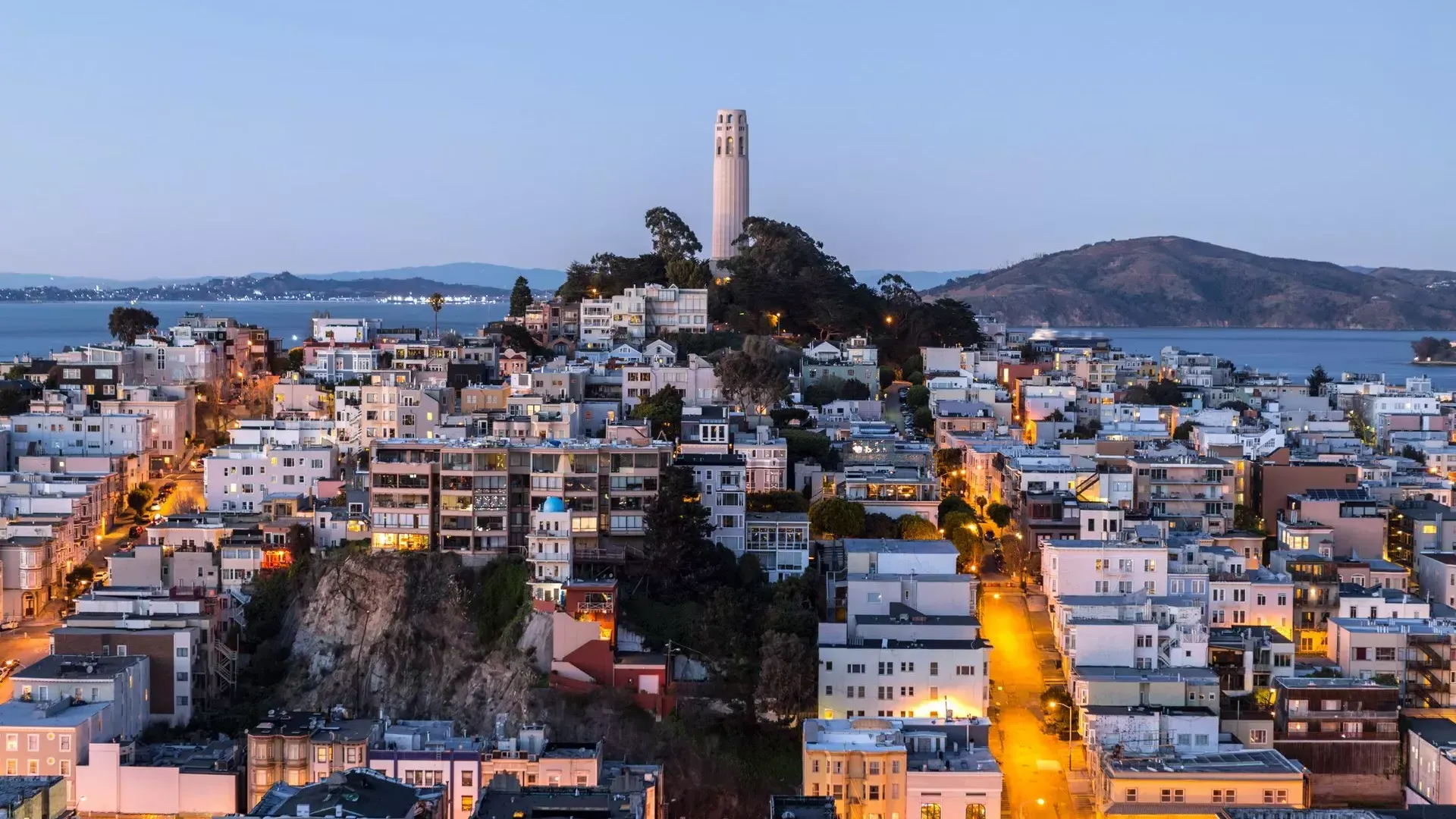 Coit Tower di San Francisco al tramonto, con le strade illuminate davanti e la baia di San Francisco alle spalle.