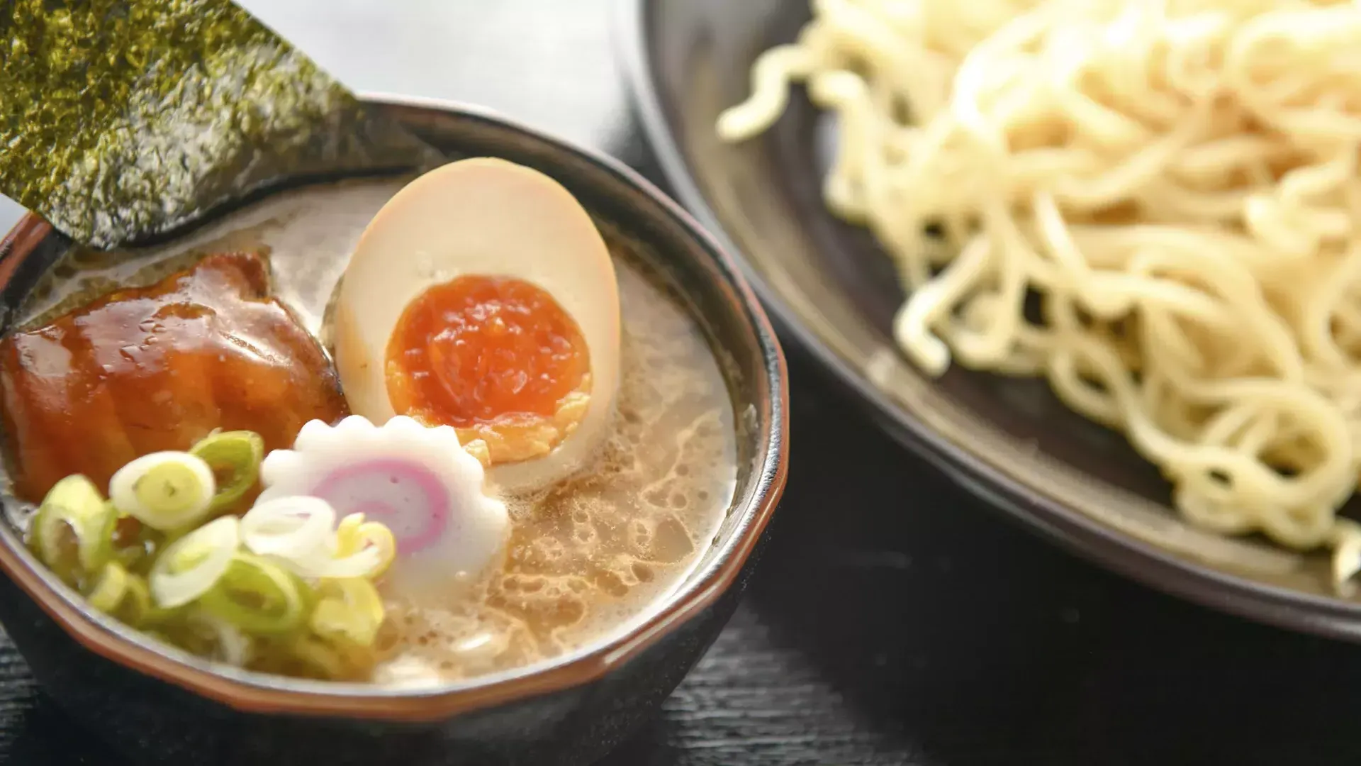 Primer plano de un plato de fideos y un plato de sopa de ramen con un huevo escalfado, cortado por la mitad.