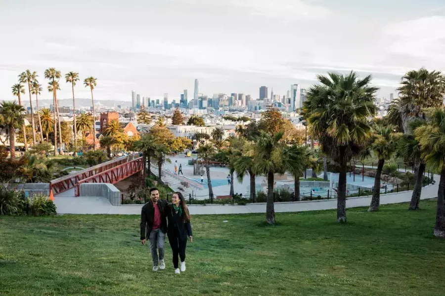 Una pareja camina hacia la cámara con Dolores Park y el horizonte de San Francisco detrás de ellos.