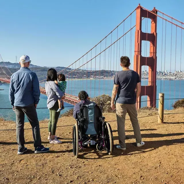 Un gruppo di persone, tra cui una persona in sedia a rotelle, è ripreso da dietro mentre osserva il Golden Gate Bridge da Marin Headlands.